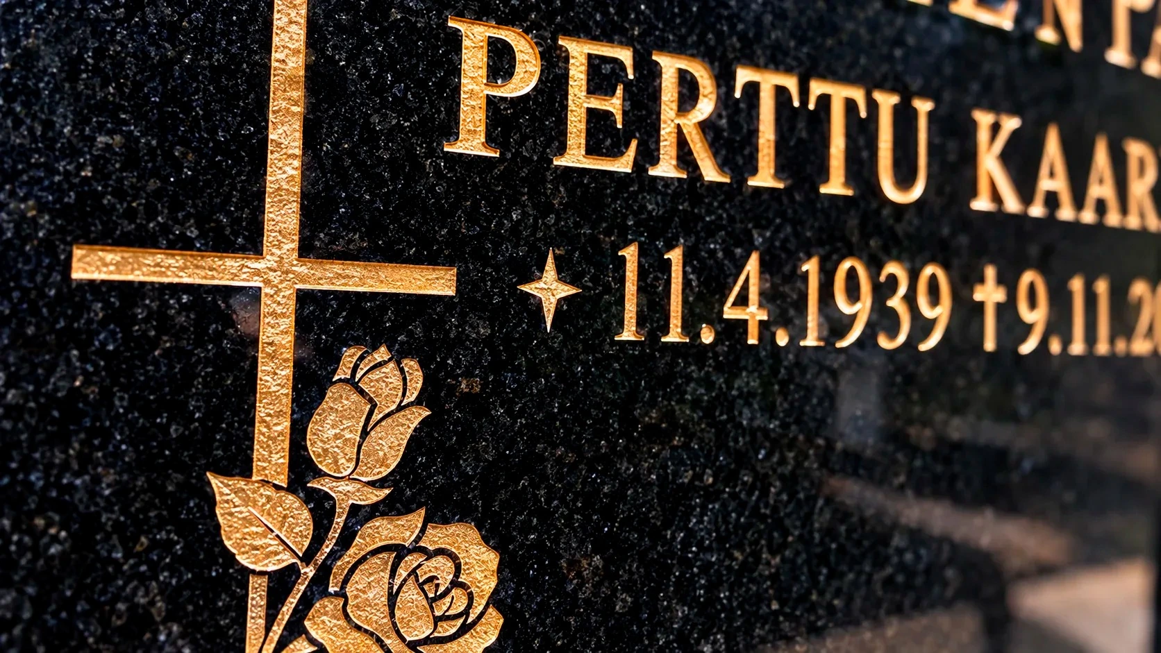 Lähikuva kullatusta hautakivikaiverruksesta mustassa graniitissa: PERTTU, risti ja ruusukoriste